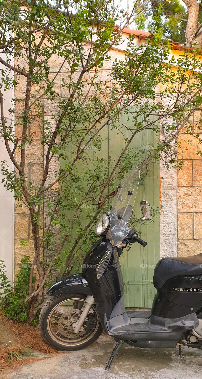 A black motobike stands near a stone house with green wooden shutters.  Summer mode of transport