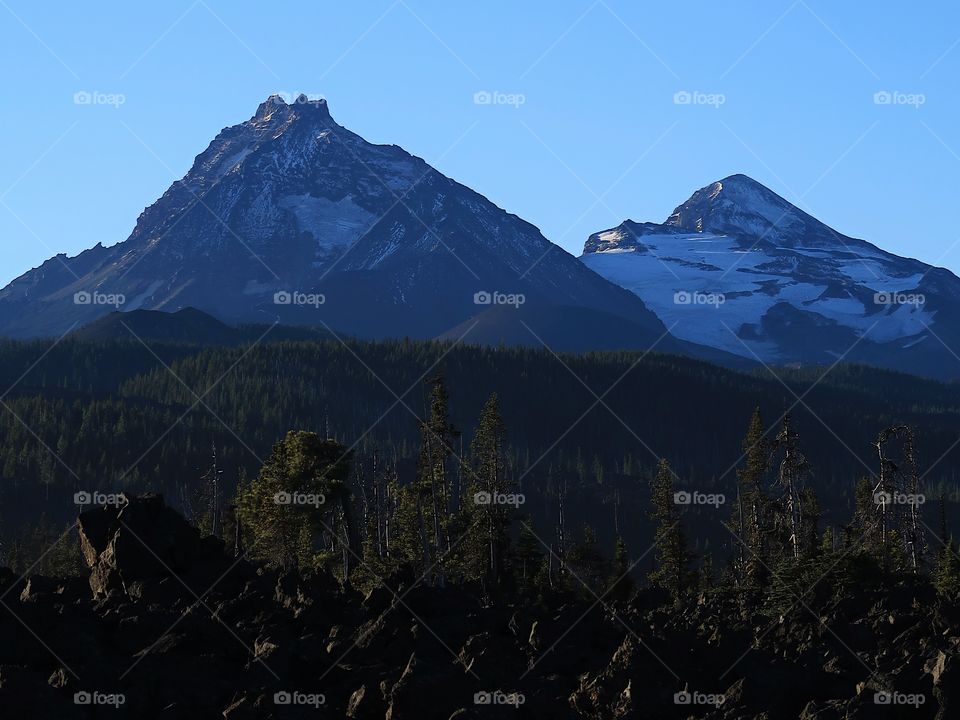 Golden morning glow of sunrise as a new day dawns on the North and Middle Sister in Oregon’s Cascade Mountain Range on a fall morning with clear blue skies.                           