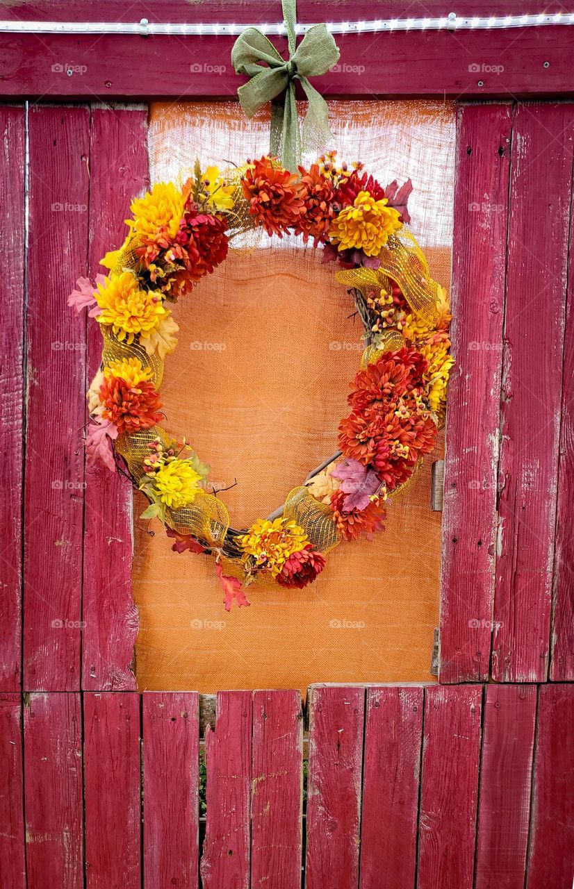 A country craft wreath hanging outdoors off of wood slats painted a barn red distressed color.