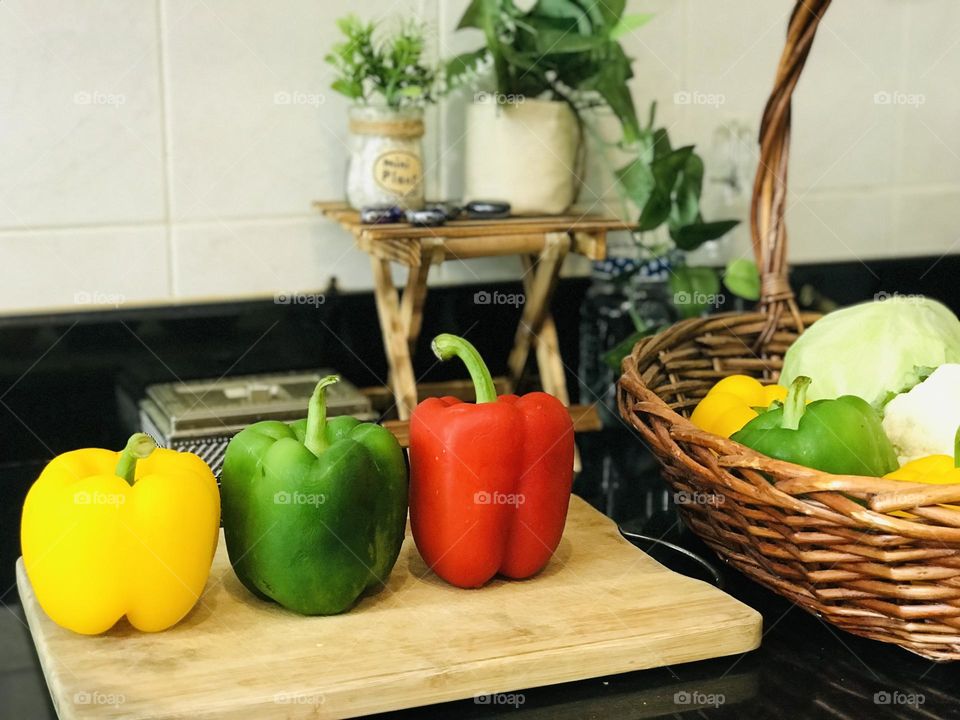 Different colours of bell pepper on wooden chopping board and vegetables in wooden bamboo basket and some mini plants on wooden small table 