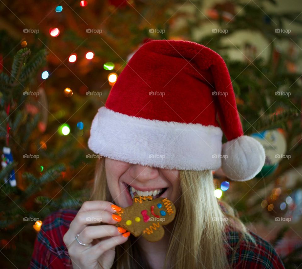 Another won’t hurt; Woman taking another bite off a Gingerbread Man in a Santa hat in front of a lit Christmas tree