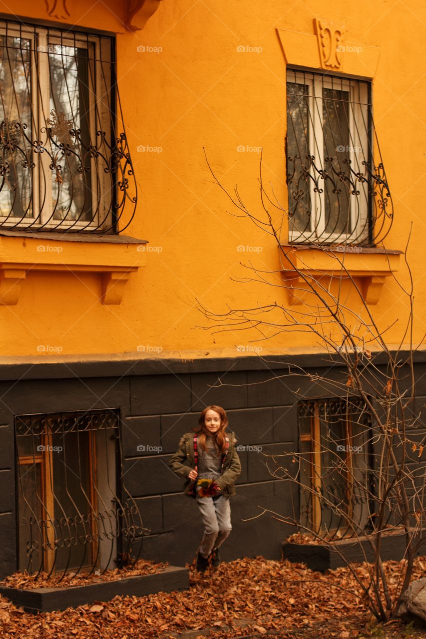Little girl jumping on a background of yellow-gray house with autumn leaves in her hands
