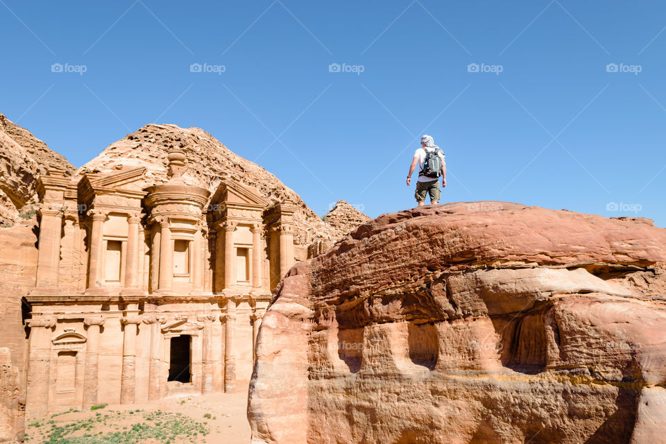 Young man traveler on the rock while observing Petra ancient monument, blue sky, travel