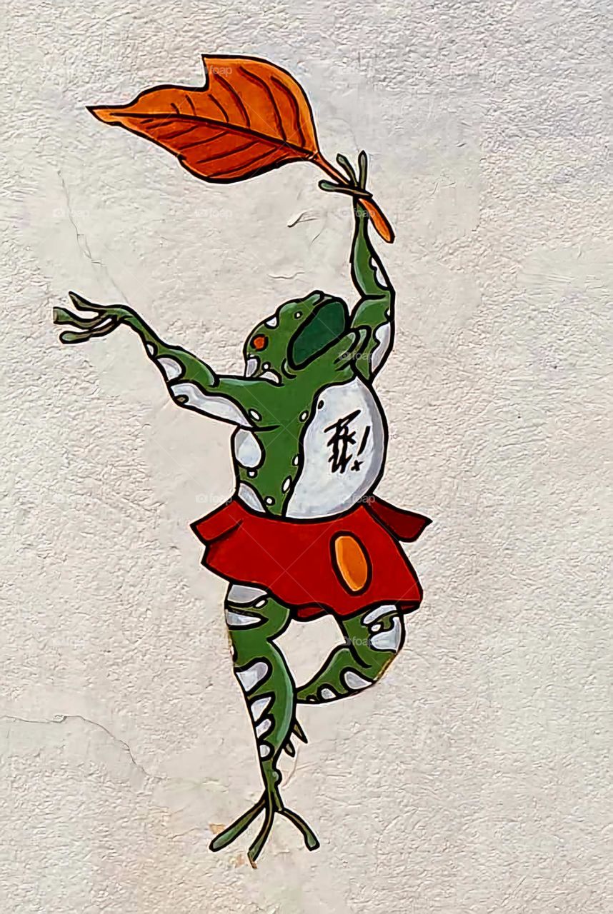 Close up on a wall art in Paris representing a green frog wearing a red skirt and waving a red leaf