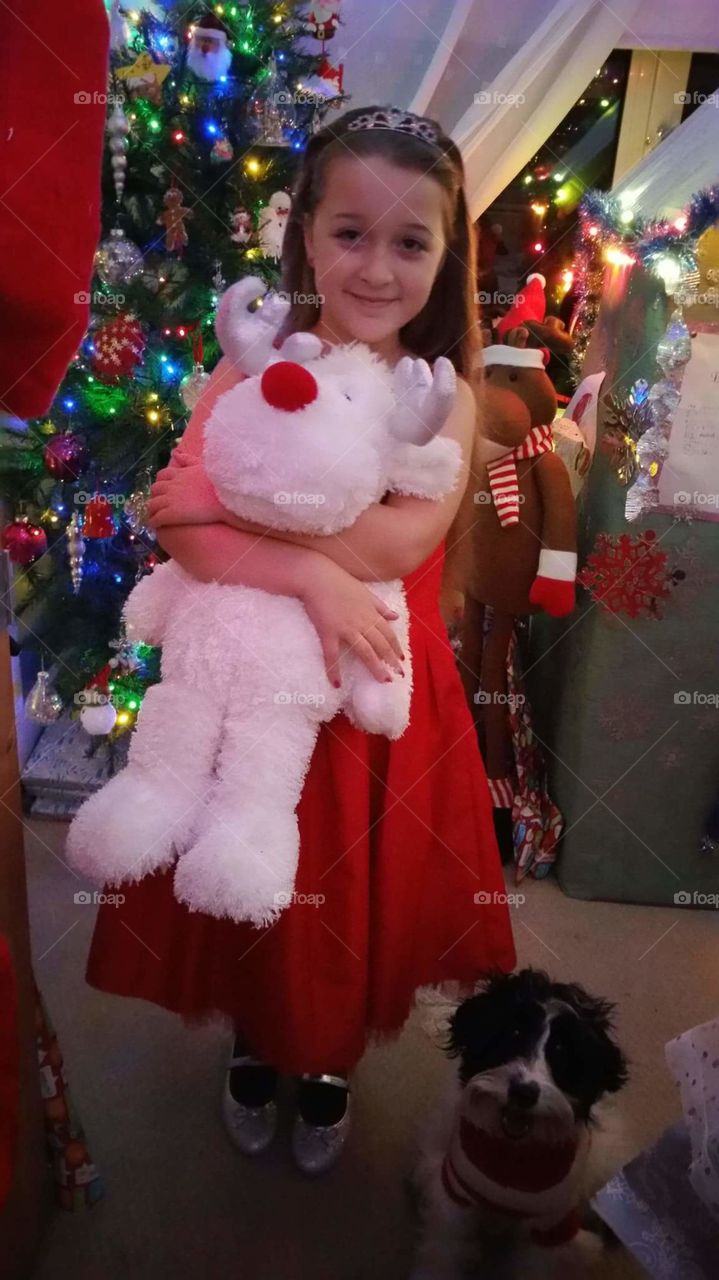 little girl holding festive reindeer toy in front of a beautiful bright Christmas tree.