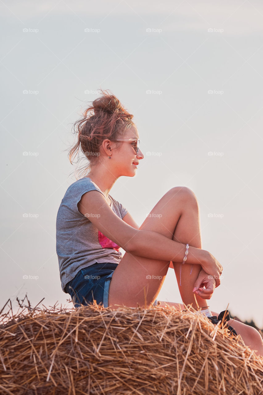 Teenage girl sitting on a hay bale at sunset relaxing while spending summertime in the village. Candid people, real moments, authentic situations