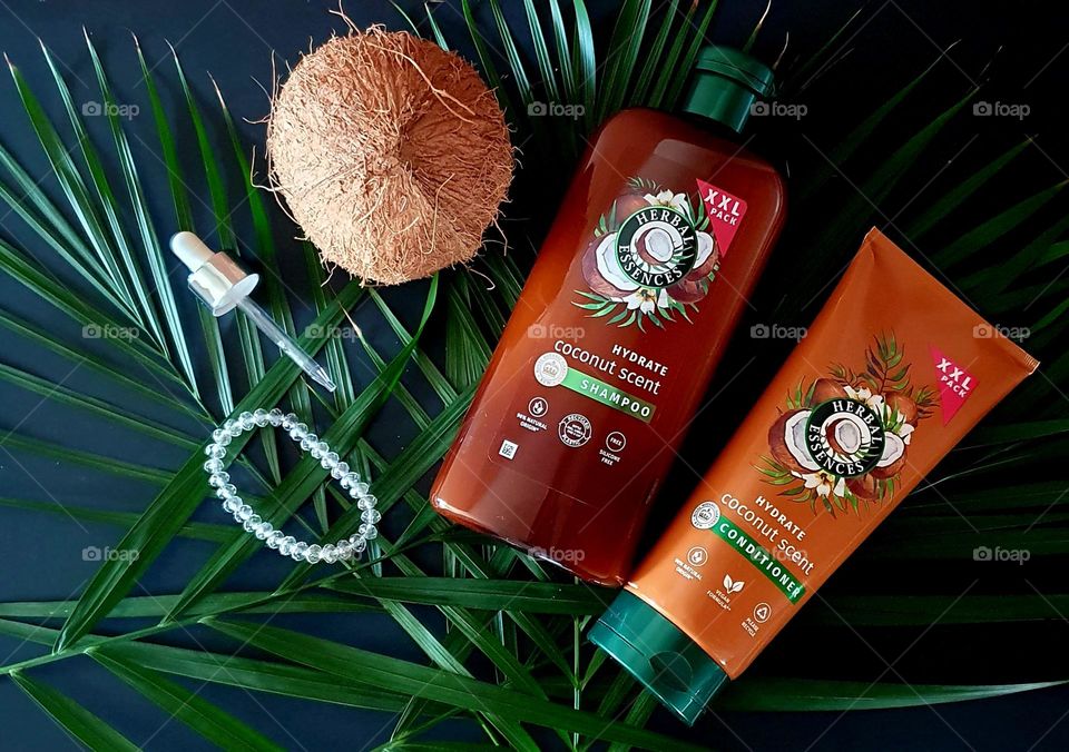 Flatlay on black paper of XXL Herbal Essences Coconut Scent shampoo and conditioner placed on palm leaves with a coconut, dropper and crystal beads bracelet