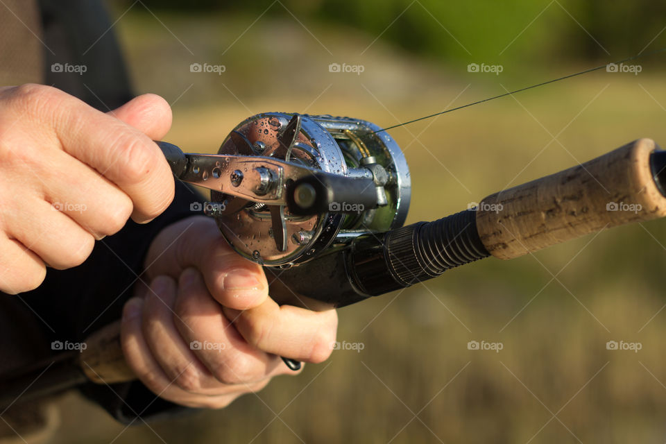 Man fishing with reel and rod. One hand on the crank and reeling fishing line in to the round reel and other hand holding fishing rod with blurred nature background.