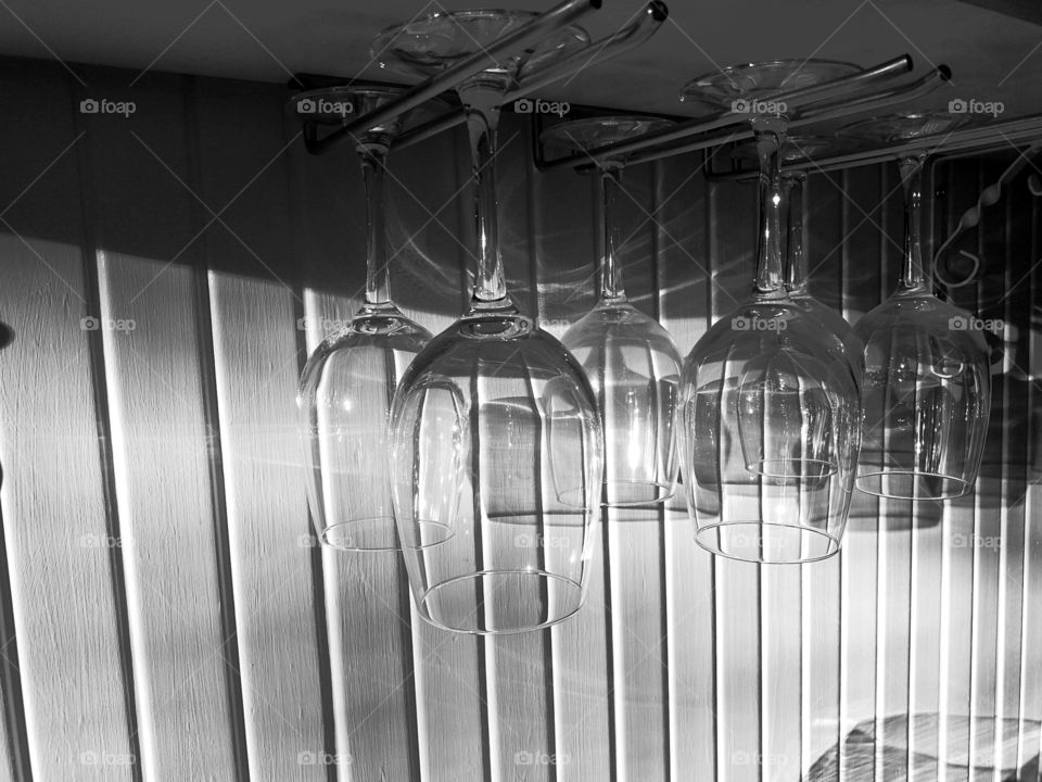 Six see-through identical wineglasses are hanging on the metallic holder upside down and the sun gives the reflection and the shadow on their surfaces and the white wall. The spiral stand under the shelf.