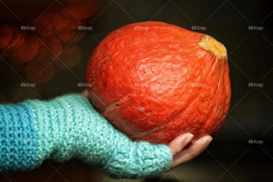 A hand wearing a blue crocheted glove holds an orange Hokkaido pumpkin against a darkly lit background