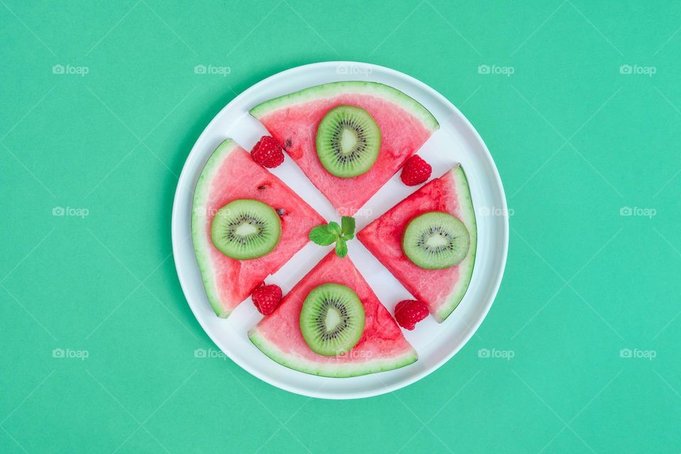 One round plate with four triangular slices of ripe watermelon and kiwi slices lie in the center on a green background, flat lay close-up. Summer treat concept.