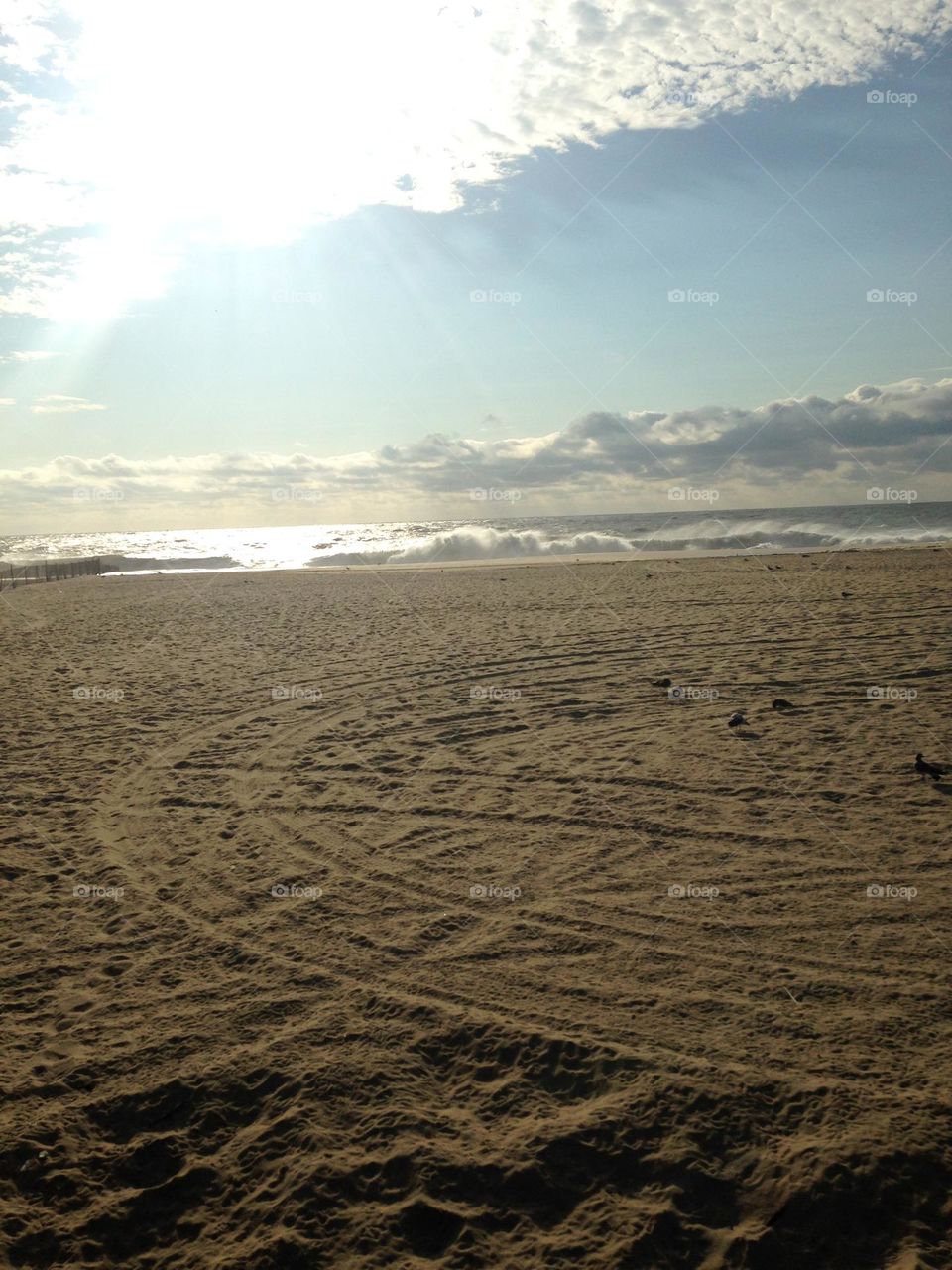 A sandy beach with circular tracks in it underneath a partly cloudy September sky. The whitecaps in the water resemble the clouds above, and a bit of sun peaks through and reflects on the water. Taken in Point Pleasant Beach, NJ. 