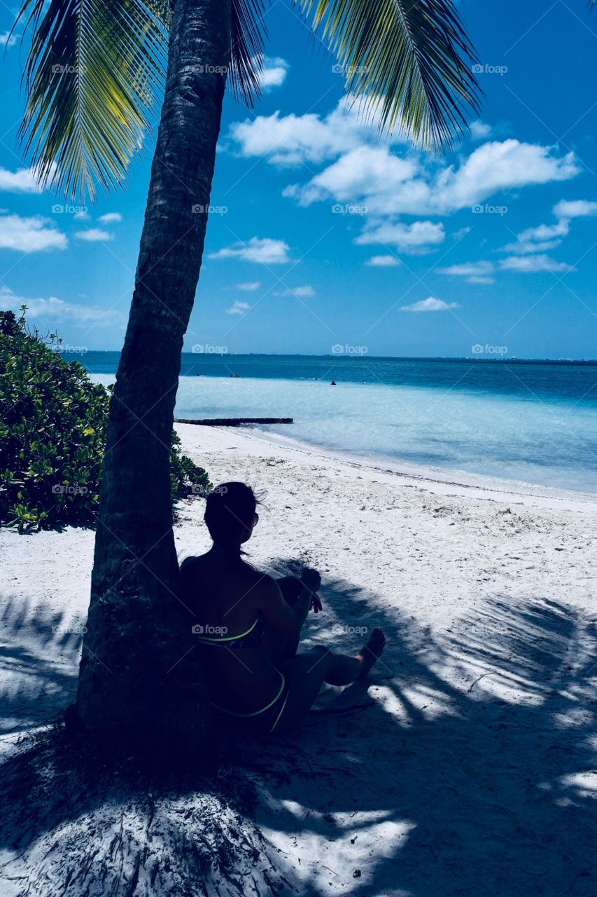 A woman sitting underneath a palm tree and watching the sea 