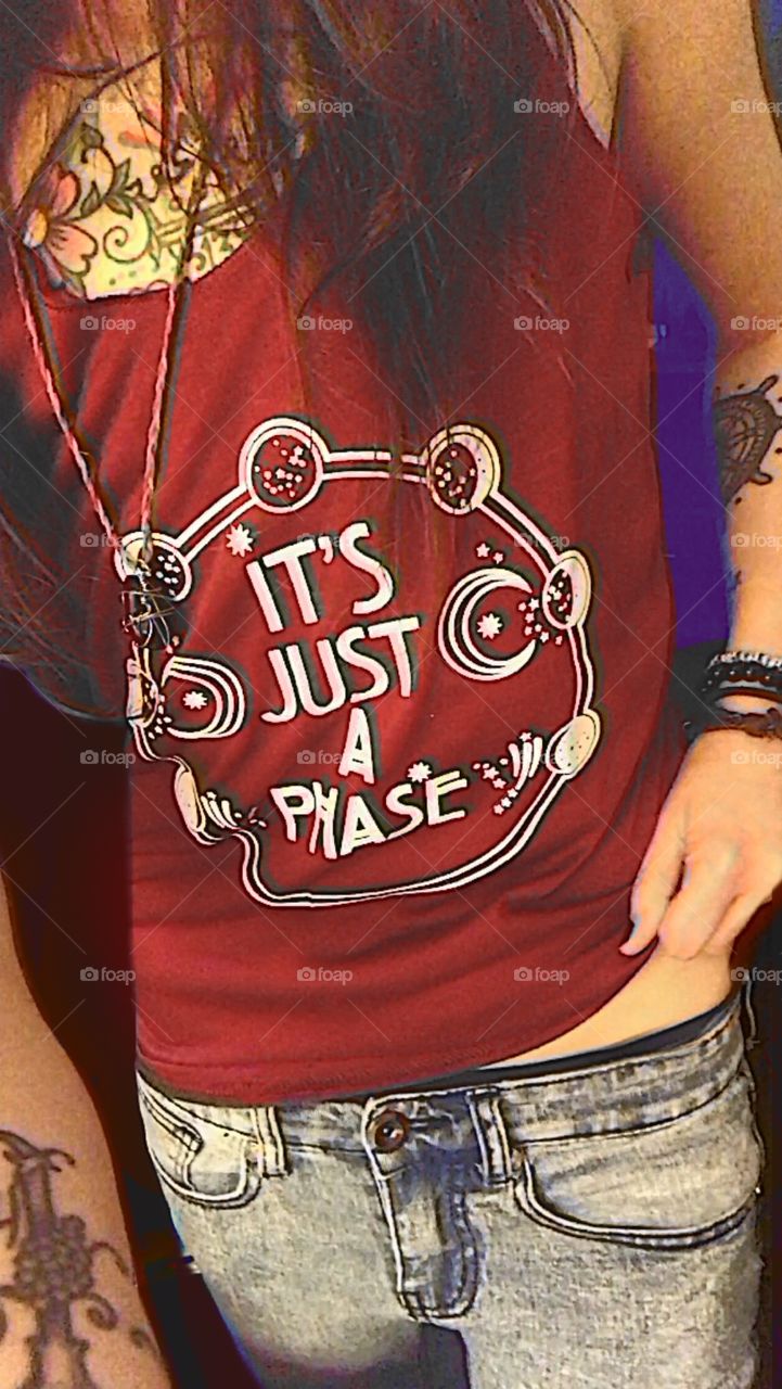 Torso of a person with tattoos lifting the corner of a red shirt with a design of a cycle of moon phases surrounding the words “It’s Just A Phase”. They have long hair and are wearing a necklace and bracelets made of stones, rope, and beads. 