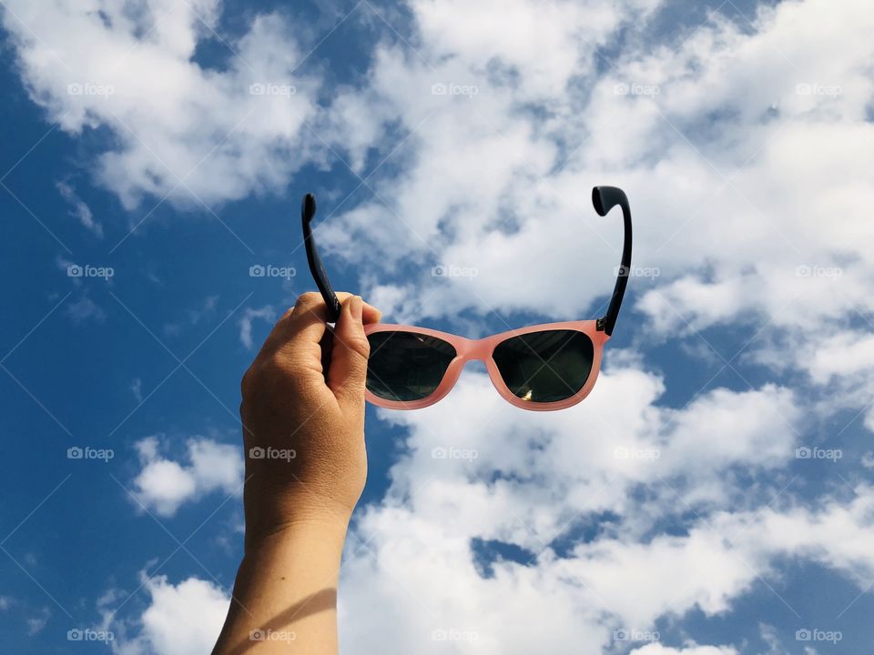 Hand holding pink sunglasses against summer blue sky covered with fluffy white clouds