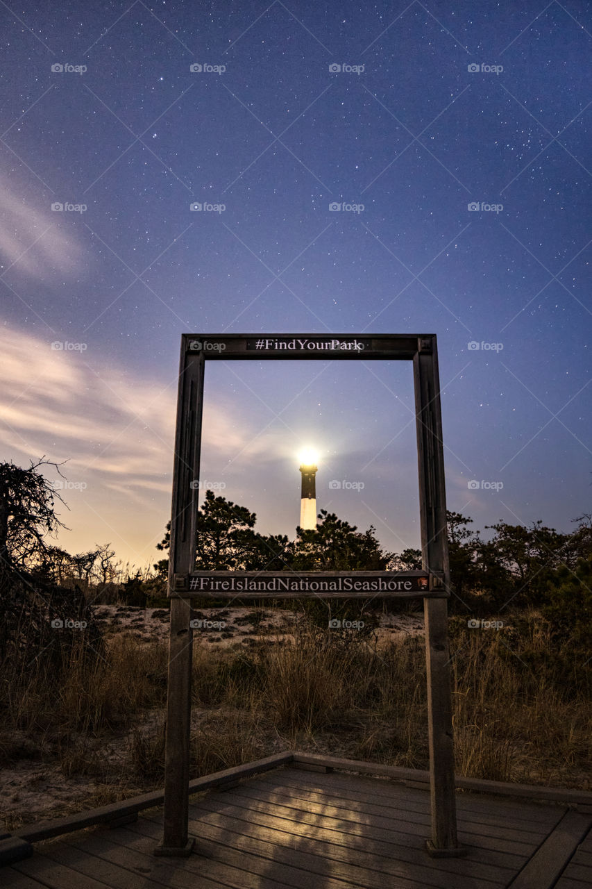 A square wooden border frames a lighthouse under a starry night sky. Fire Island National Seashore