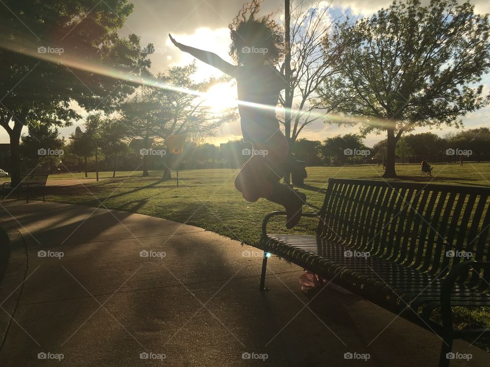 a girl jumping from a bench in a park during the sunset