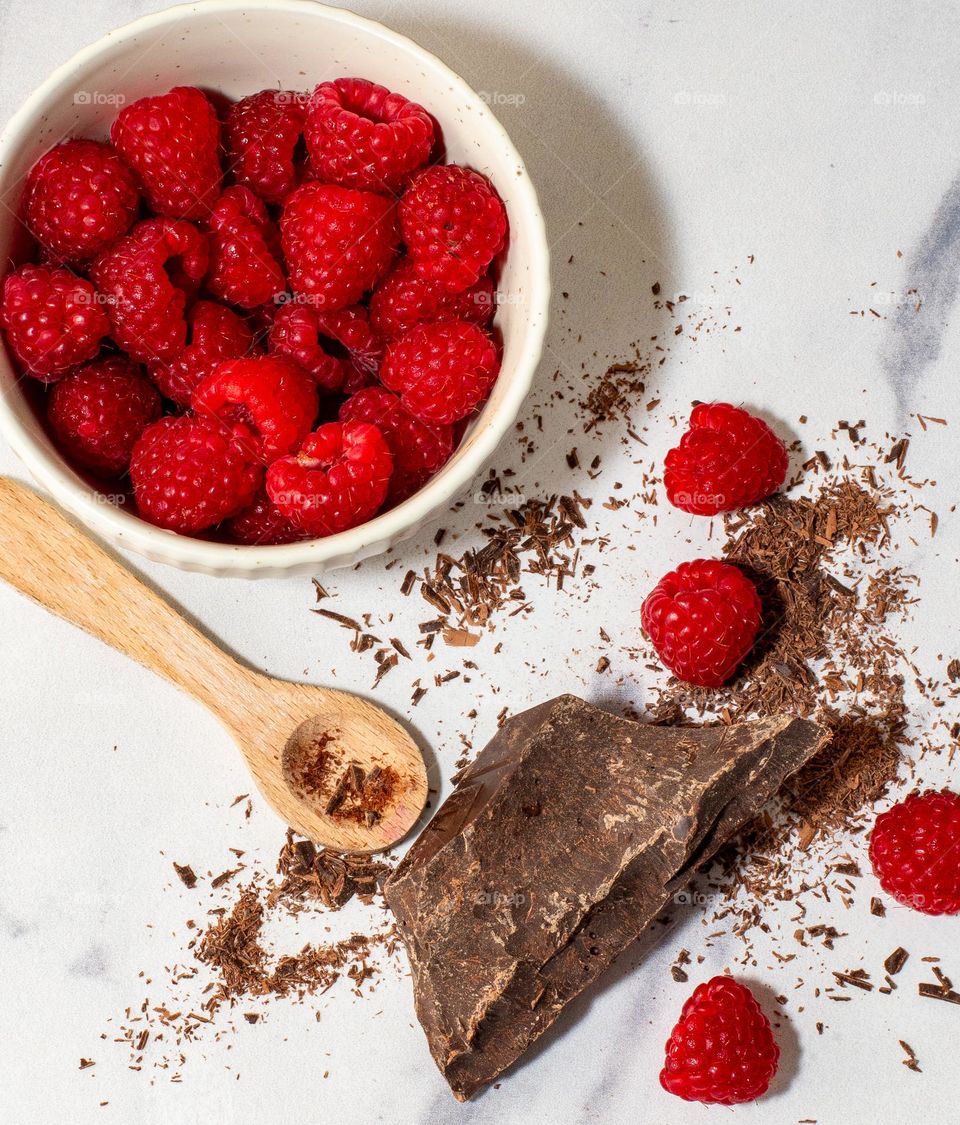 Chocolate chunks and raspberries in a bowl on a marble background with a small wooden spoon