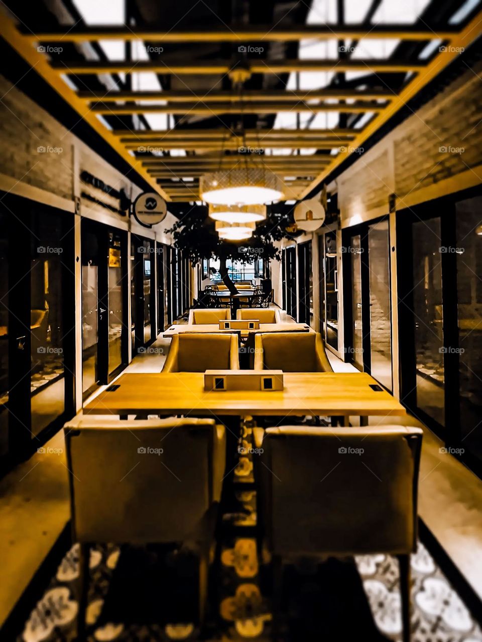 Portrait of the interior of a long room with neatly arranged tables and chairs, surrounded by glass walls, and equipped with lighting from hanging lamps