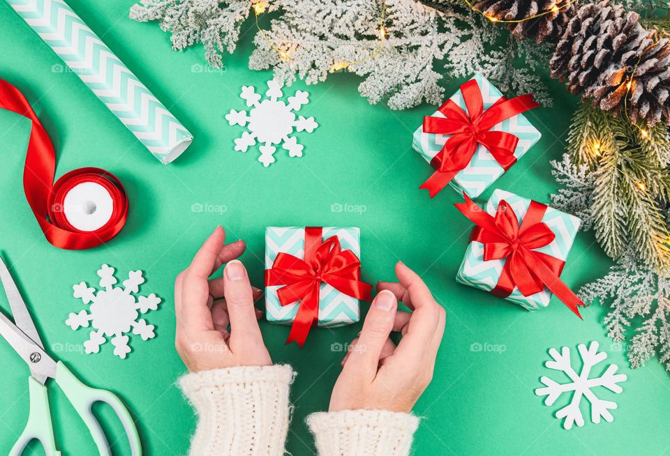 The hands of a young caucasian girl in a white knitted sweater hold a gently small gift box with a ribbon, and around are decorative snowflakes, scissors, a spool of red ribbon, a roll of wrapping paper and a fir branch with a garland, flat lay close