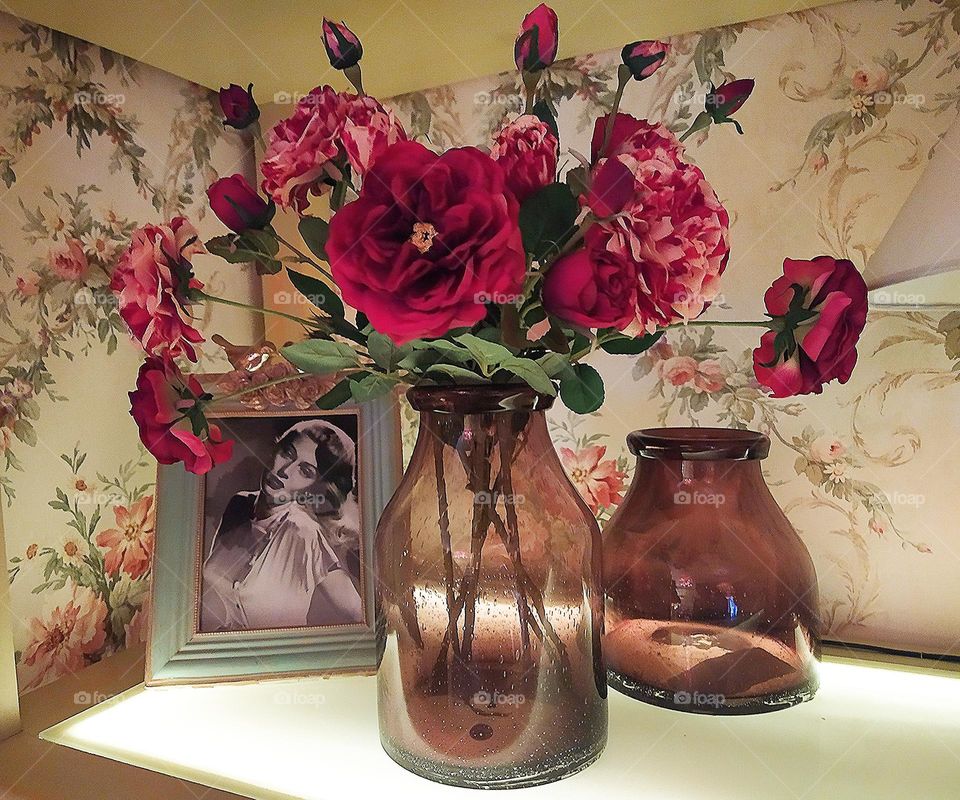 There are dark glass bottles on the shelf. In one bottle there are roses in the form of blossoming flowers and buds. Roses in all shades of red. Green leaves break through between the roses. In the background is a framed black and white photograp
