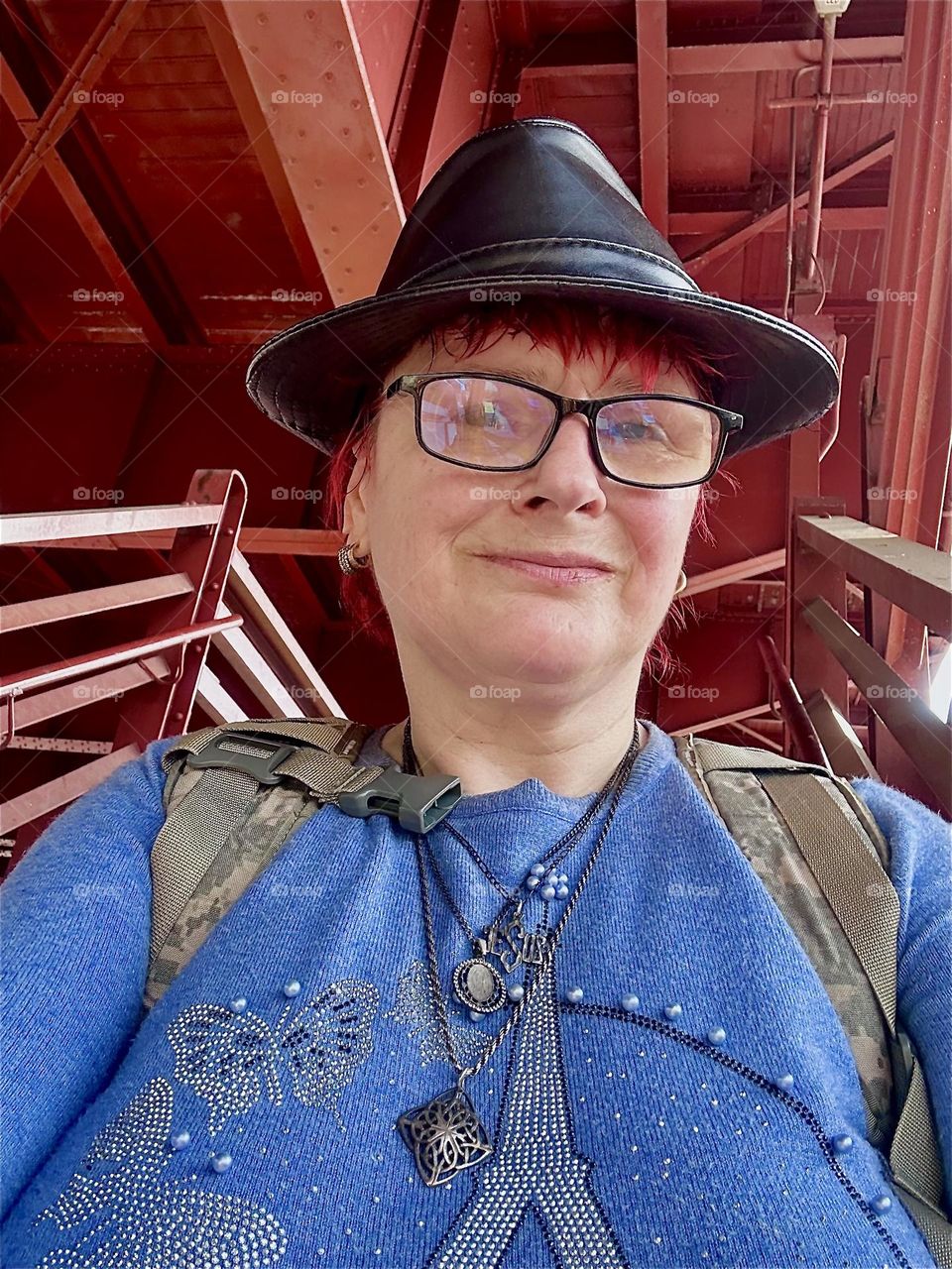 This is me, “Petra Schiller” a.k.a. “Maxine Firefox” on the red metal staircase of the “Pulaski Bridge” in LIC, Queens. My light blue sparkly sweater is complementary to the orange red of the Bauhaus architecture behind me. 2025. Hypnotic Productions