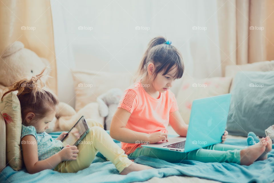 Little sisters with gadgets(laptop and tablet) in the bed.