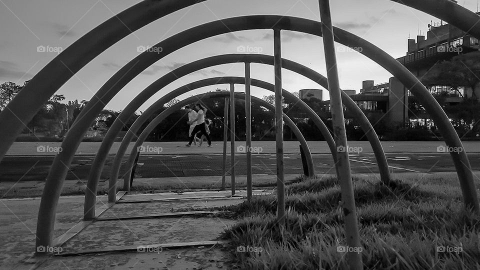 B&W photo framing through the iron arches of the bicycle parking.  In the background, family walking in the park.