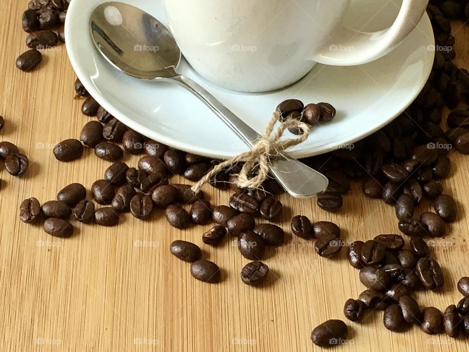 Roasted whole organic coffee beans spilled on light oak wood table surrounding white coffee cup and sauce, spoon tied with natural twine jute bow. Natural, simplicity, copy space 