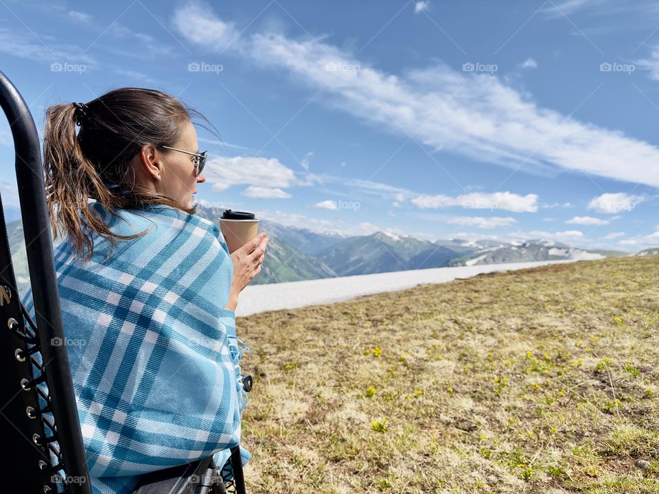 girl in a blanket with a cup of coffee sits in a chair in the mountains