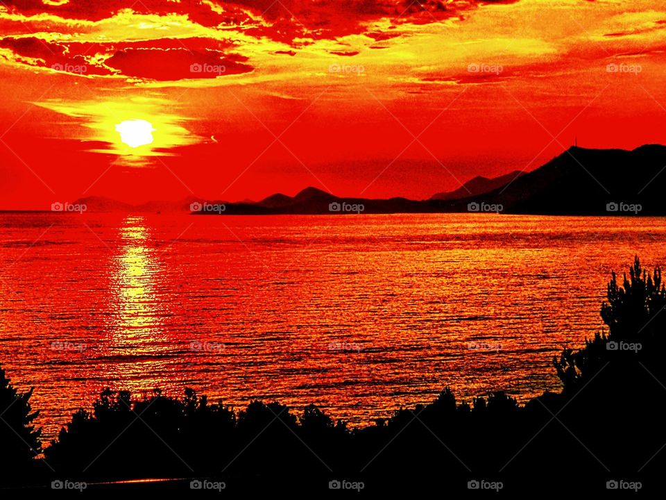 Vivid red sunset with yellow sun reflecting across the sea and edging the clouds with mountains in the background