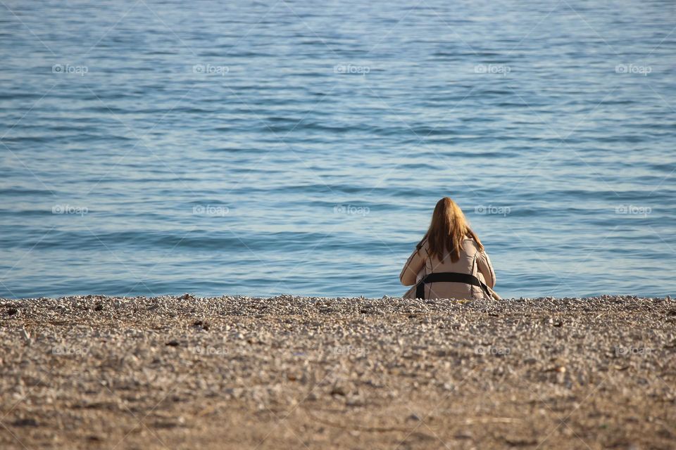 A young woman is sitting on an empty beach wearing a warm jacket