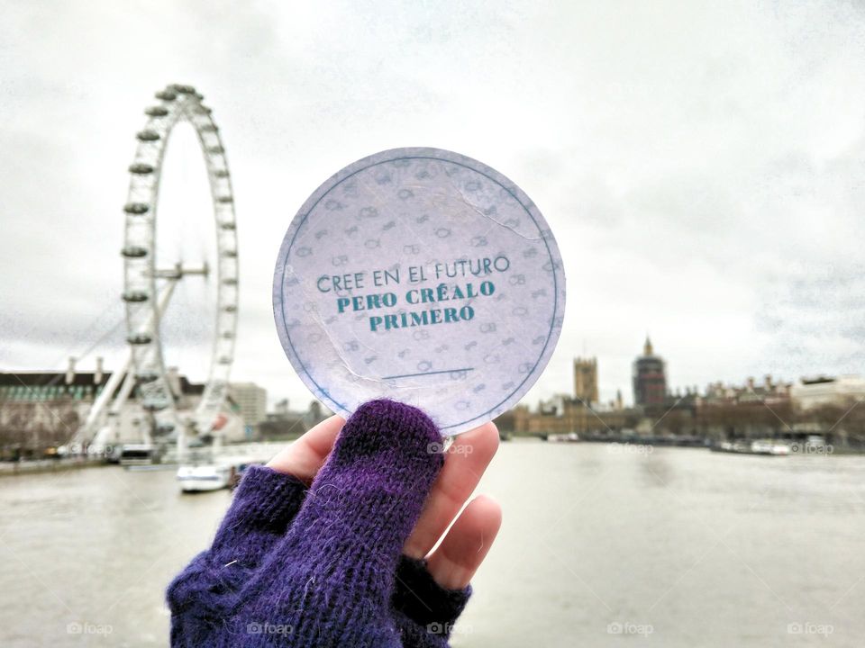 landscape of the city of london with the river thames, the london eye and big ben, and in the foreground a sign that says "believe in the future but create it first" held by a gloved hand.