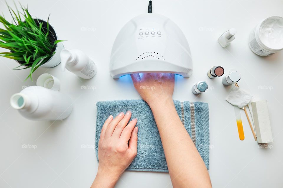 Woman applying gel hybrid polish using UV lamp. Beauty wellness spa treatment concept. Cosmetic products, UV lamp, green leaves on white table. Spa, manicure, skin care concept. Flat lay, overhead view