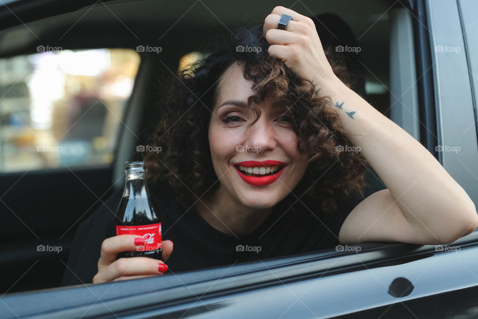 beautiful curly-haired brunette sitting in the car and drinking Coca-Cola, a delicious drink.  the girl is going to go to work. Happy smiling girl 