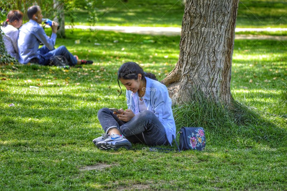 a girl in jeans and a shirt sits on a lawn in a park with a phone in her hands