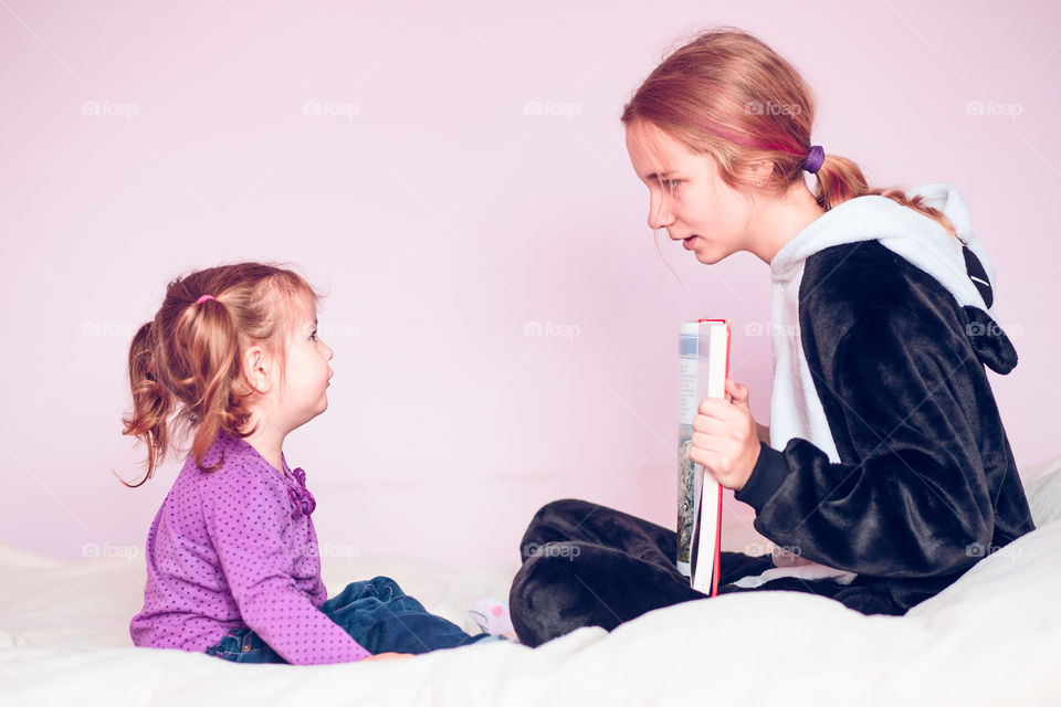 Girl showing the pictures in a book her younger sister while sitting on bed at home