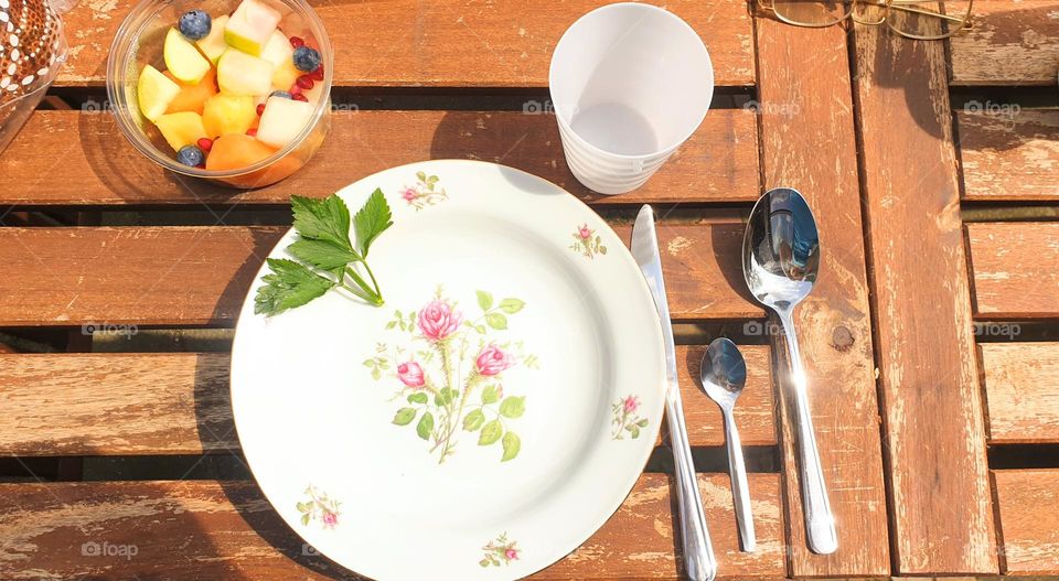 time for outdoor lunch with a porcelain plate with roses herbs fruit cutlery and a white cup on a wooden table