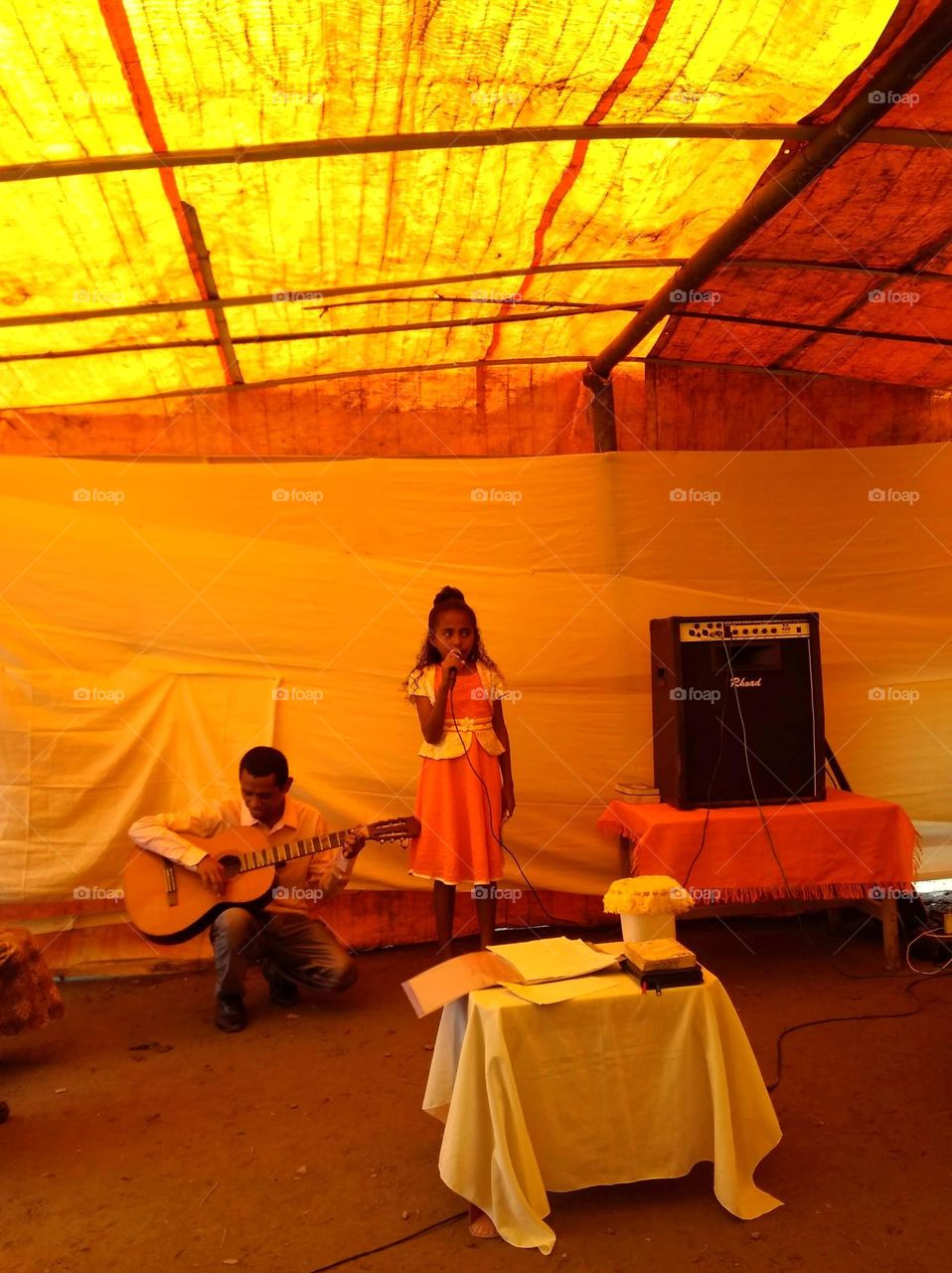 A performance of a girl singing and a man playing guitar under a tent made of orange and yellow plastic. Orange hues due to daylight