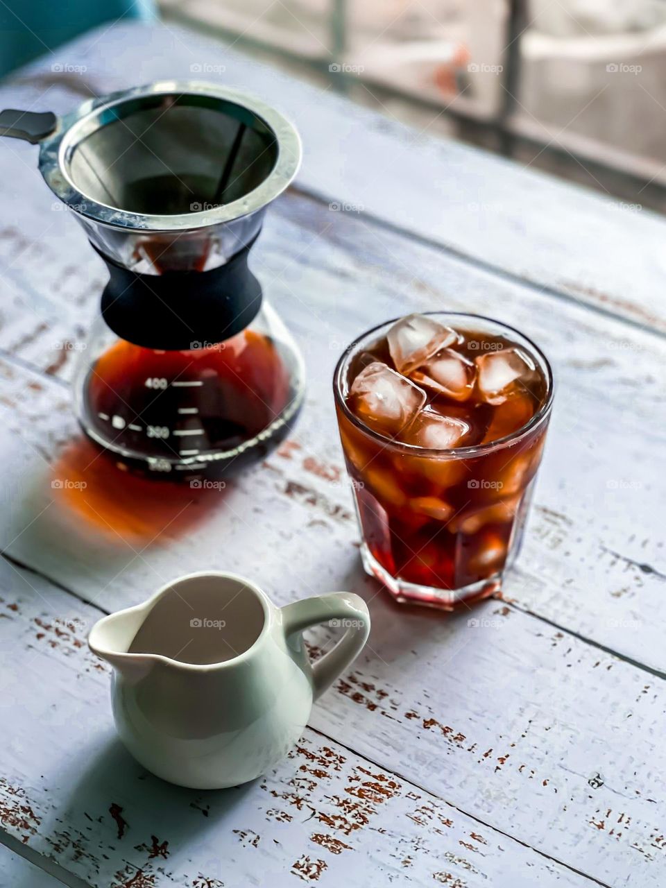 A cup of black coffee ice with coffee dripper at the side and milk jar at the front on white wooden table