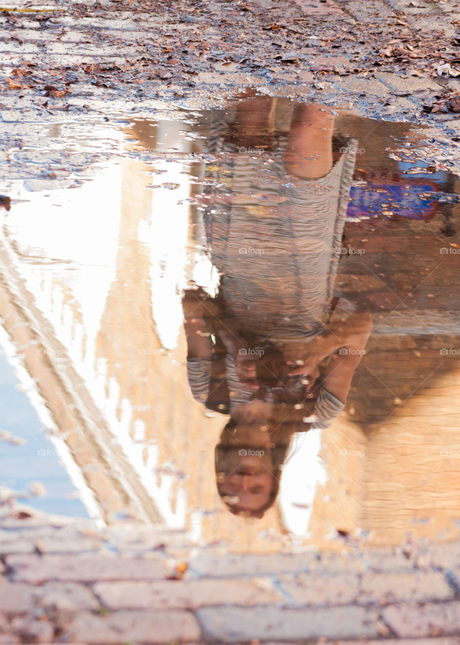 image of a girl with a camera in front of a tan and white building all reflected upside  down in a puddle on a brick road