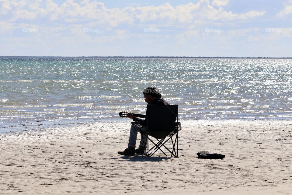 Guitarist playing guitar while sitting on chair on beach surrounded by glistening ocean at the golden hour late day