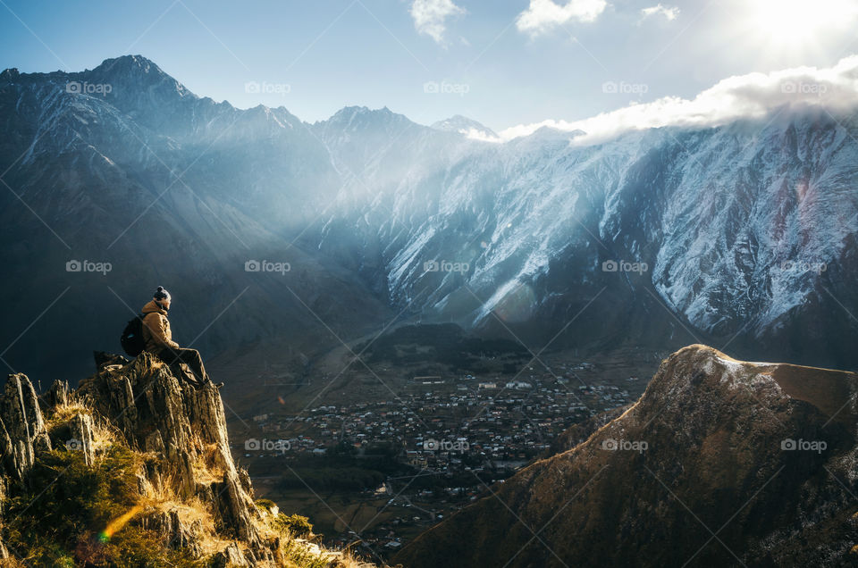 Young tourist with backpack sits on the cliff and looks at the misty mountain village and glacier at sunrise, Stepantsminda, Caucasus, Georgia