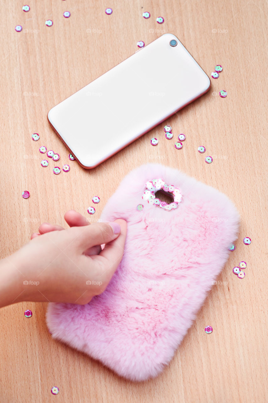 Close shot of female hand decorating phone case with sequins following diy ideas on wooden table