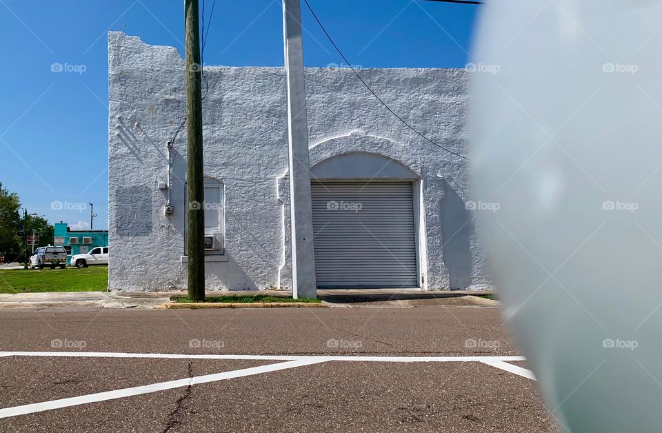 Old white architectural building with signs of aging elements with arched garage door under a blue sky in the afternoon, seen from the other side of the street from a rounded top white pole.