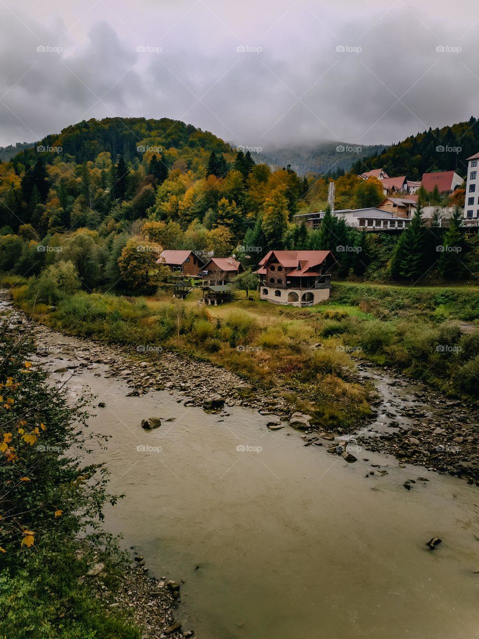 Colorful autumn forest in the mountains, village behind the river, foggy weather