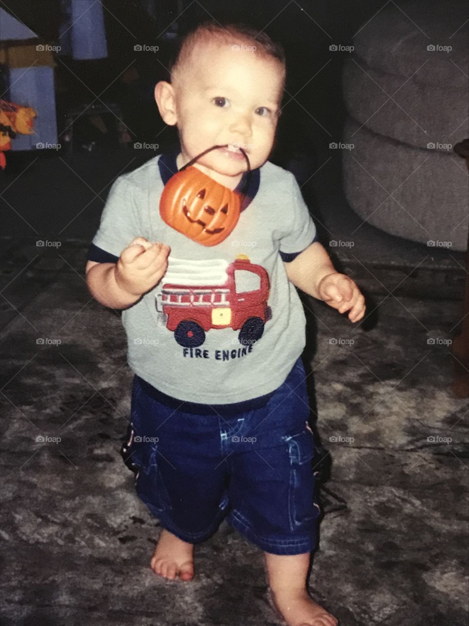 Toddler boy wearing a fire engine shirt and holding a little pumpkin bucket with his teeth.