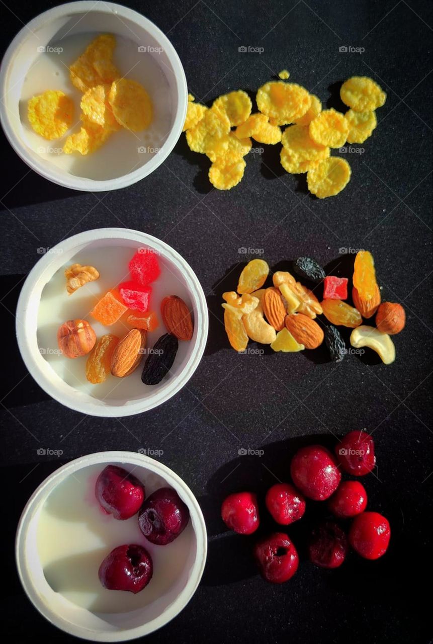 Food. On a black background are three jars of white pudding.On the surface of each pudding are different ingredients: nut mix,corn flakes and frozen cherries. Ingredients are scattered around each pudding jar:nut mix,corn flakes and frozen cherries