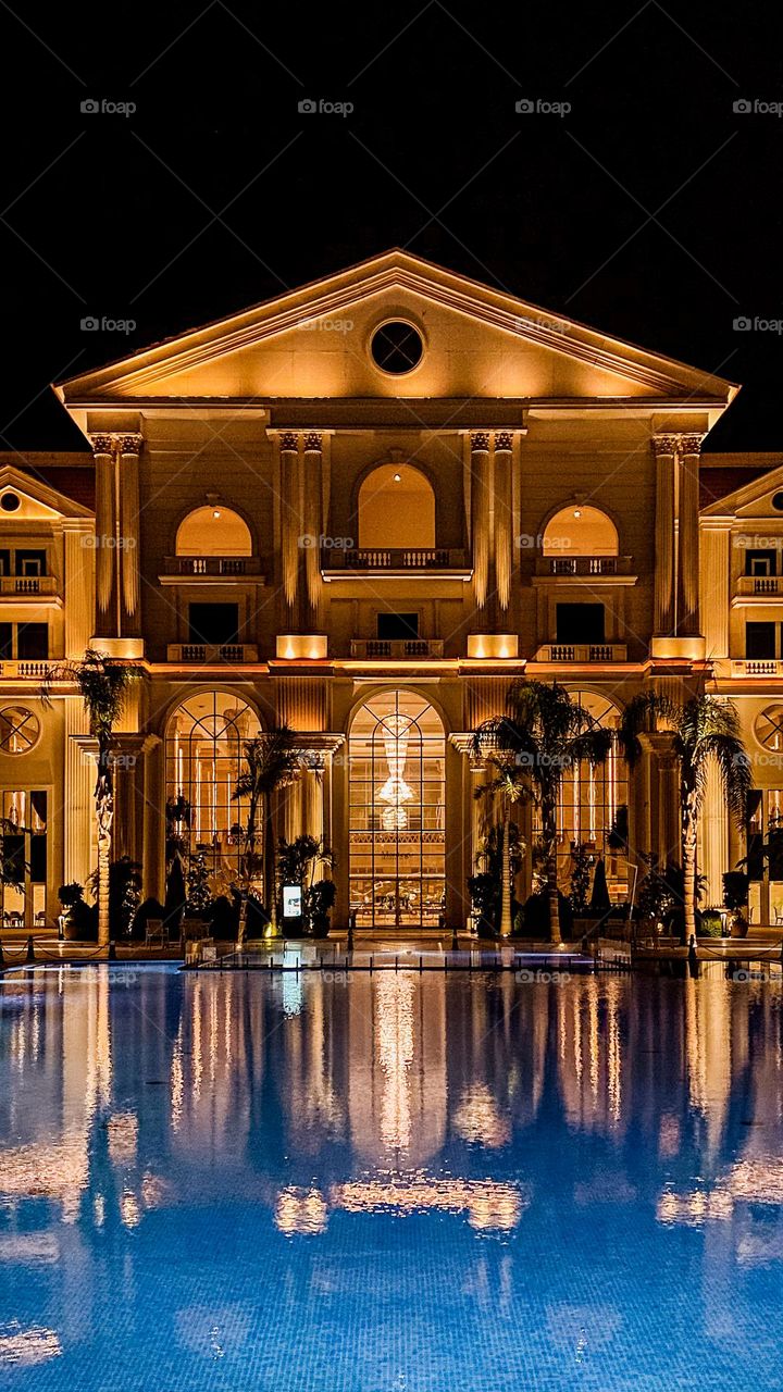 Night view of a mansion with lit up windows and reflections on a swimming pool, evoking a sense of warmth and grandeur.