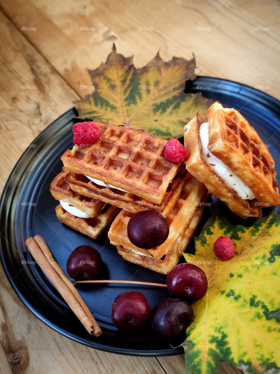 Composition. On a wooden background. A black plate containing Belgian waffles stuffed with whipped cream. Red cherries, cinnamon sticks, raspberries and dried autumn leaves